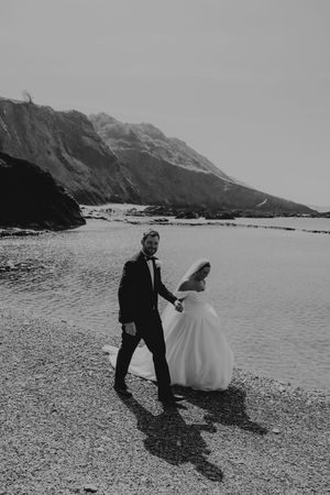 Black and white photo of bride and groom walking hand in hand along a rocky beach with dramatic cliffs.