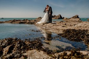 Bride and groom share a romantic kiss on rocky coastal shoreline with turquoise ocean waters and clear blue sky.