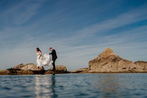 Bride and groom walk on coastal rocks with flowing white dress, blue sky and calm ocean waters in background.