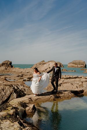 Bride and groom walk hand in hand across rocky coastal terrain with turquoise ocean backdrop.