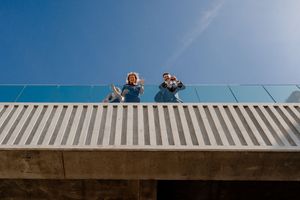Two people waving and smiling from a modern rooftop balcony with glass railing against a clear blue sky.