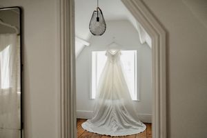 Elegant white wedding gown hanging in a bright white room with wooden floors and pendant light.