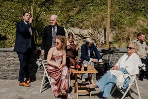 Wedding guests socializing outdoors on a sunny patio, with women in elegant dresses and men in suits enjoying drinks.