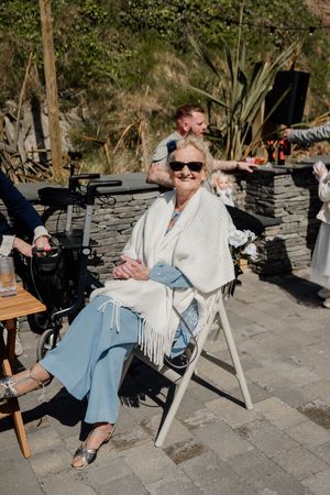 Elderly woman smiling in sunglasses and white shawl seated at outdoor wedding reception on sunny day.