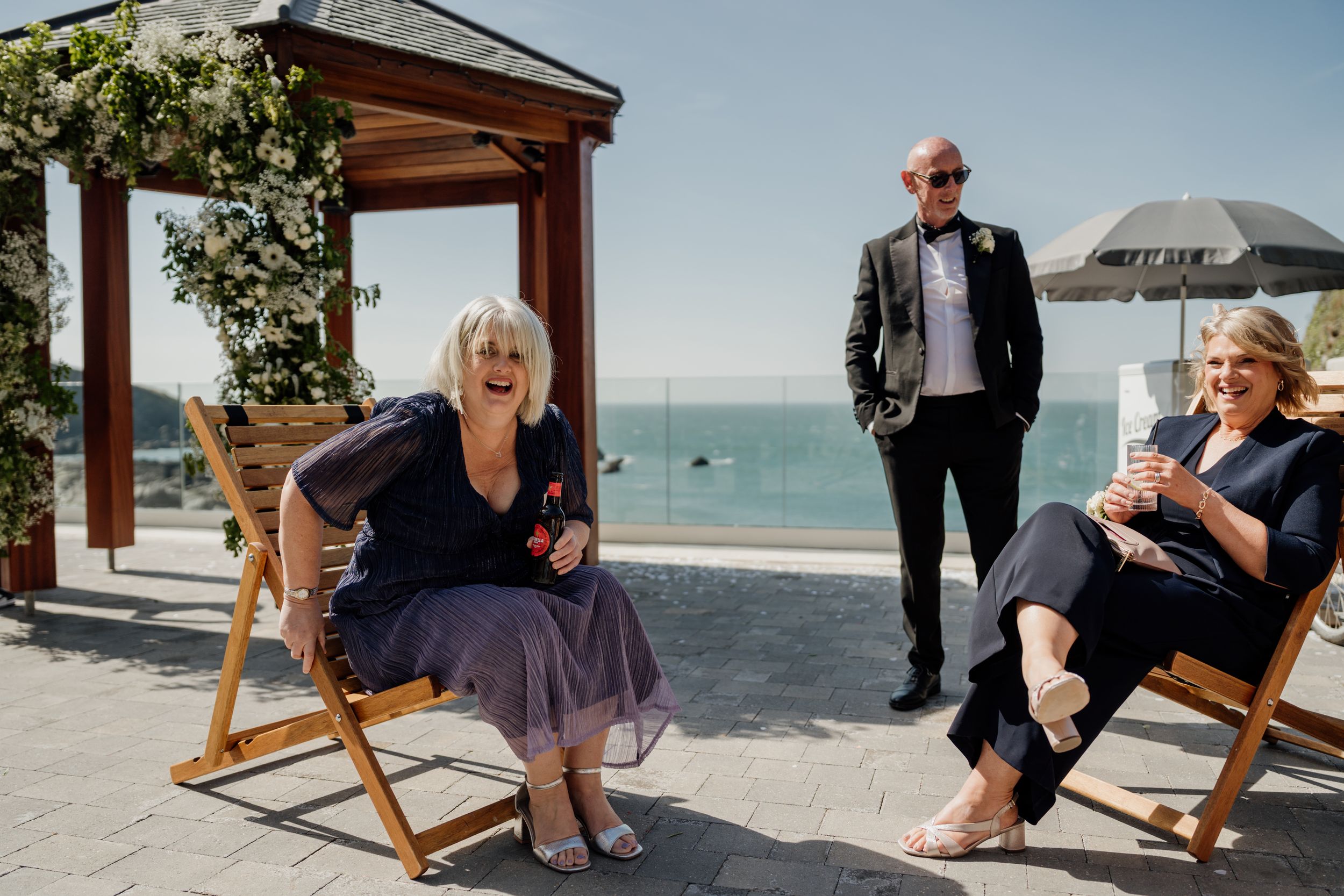 Three elegantly dressed wedding guests relaxing on wooden deck chairs by the seaside at a coastal venue.