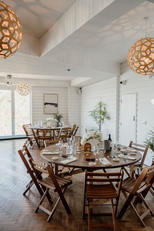 Elegant wedding reception venue with wooden round tables, folding chairs, herringbone floors, and geometric pendant lights.