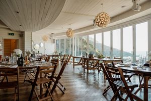 Elegant wedding reception venue with wooden chairs, long dining tables, pendant lights, and panoramic ocean views.