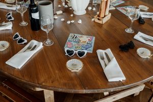Elegant round wooden wedding reception table set with white napkins, wine glasses, sunglasses, and decorative stickers.