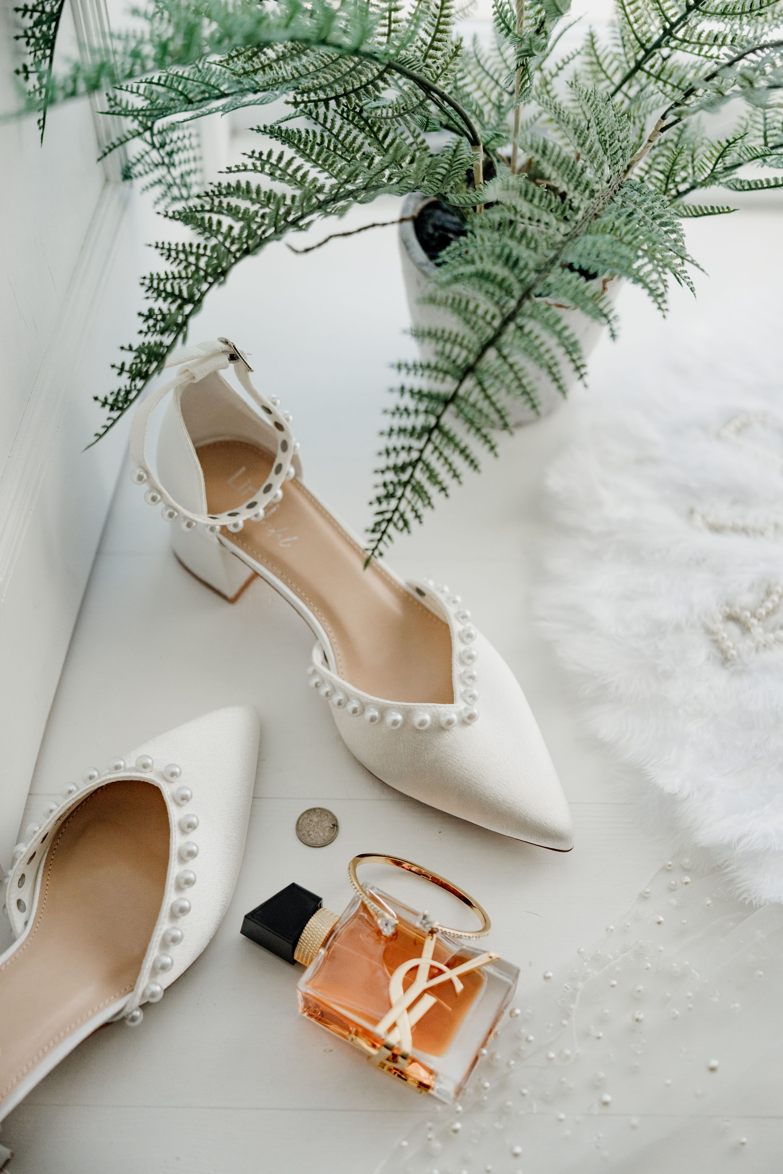 White pearl-embellished bridal heels displayed beside YSL perfume and fern greenery on a white surface.
