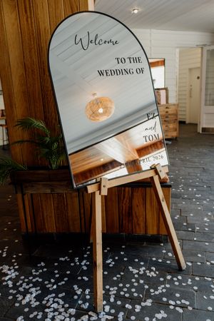 Elegant arched mirror wedding welcome sign on wooden easel with white flower petals scattered on dark tile floor.