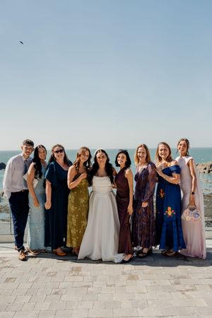 Bride in white gown poses with wedding guests in colorful dresses by the ocean on a sunny day.