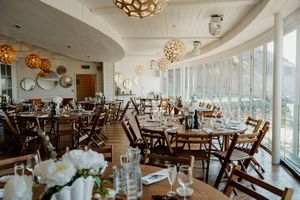 Elegant wedding reception venue with wooden chairs, white floral centerpieces, and decorative pendant lights overlooking large windows.