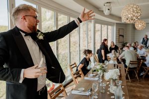 Groom in black tuxedo giving a speech at wedding reception in a bright sunlit dining room.