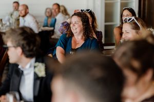 Wedding guests laughing and smiling during indoor reception speeches at a bright venue.