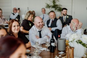 Wedding guests laugh and smile during reception speeches at elegantly decorated dining tables.