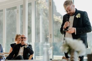 A bearded groomsman gives a speech at a wedding reception while the happy couple laughs in the background.