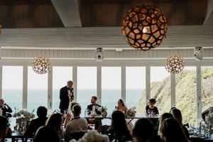Wedding speech moment with couple seated at head table, framed by ocean views and geometric gold pendant lights.