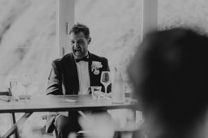 Black and white candid photo of a laughing groom seated at a table during wedding reception.