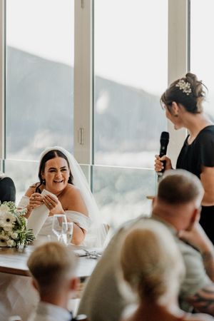 Bride laughing emotionally during a heartfelt wedding speech by a bridesmaid holding a microphone.