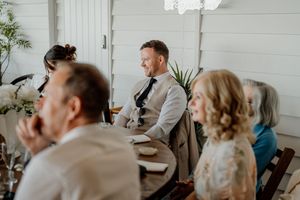 Groom in suit smiling while seated at wedding reception table with guests in bright windowed venue.