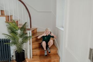 Young blonde child sitting on wooden staircase steps near a potted palm plant indoors.