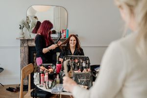 Woman smiling in mirror reflection during bridal hair styling session with makeup artist.