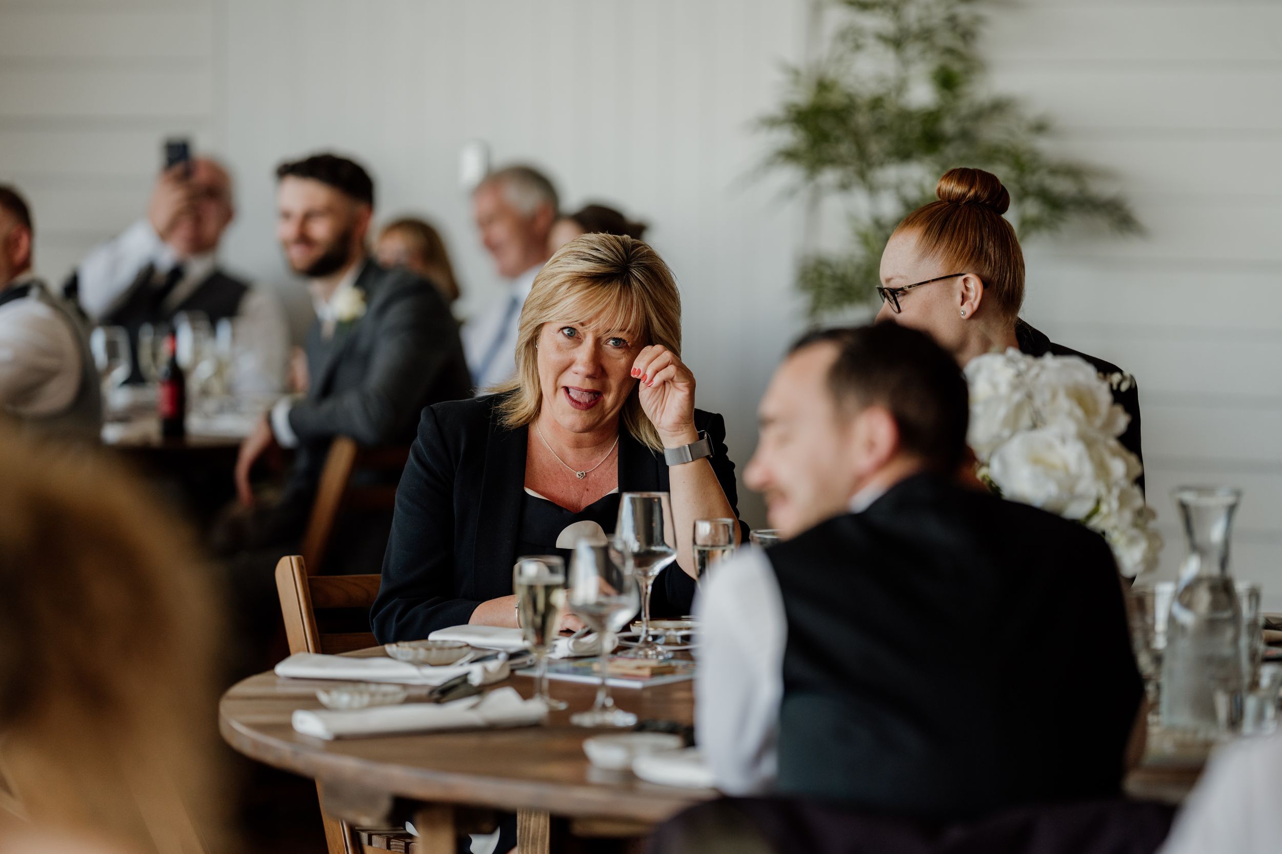 Woman in black jacket smiling at a wedding reception dinner table with guests in background.