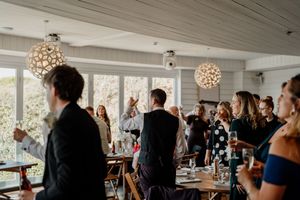 Wedding guests raising glasses in a toast inside a bright venue with decorative pendant lighting.