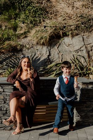 Woman in brown dress and young boy in suit enjoying ice cream cones together outdoors at a wedding.