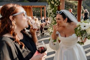 Bride in white off-shoulder gown laughing with guest holding red wine glass at outdoor wedding reception.