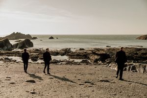 Three men in dark suits standing on a rocky beach gazing at the ocean horizon.