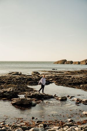 Person walking on rocky coastal shoreline with calm ocean and clear sky backdrop.