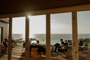 Two people relaxing on a deck with stunning ocean sunset views through large glass sliding doors.