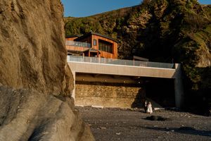 A bride and groom stand on a rocky beach beneath a modern wooden building at golden hour.