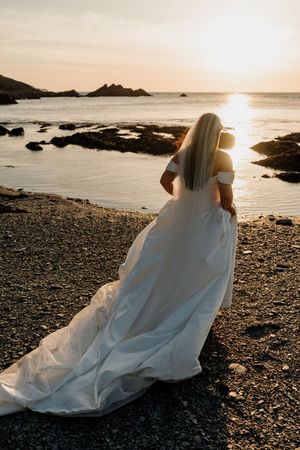 Bride in white off-shoulder gown with long train stands on rocky beach at golden sunset, gazing at the ocean.