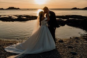 Bride and groom sharing a romantic sunset kiss on a rocky beach with golden ocean reflections.