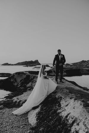 Black and white photo of bride and groom standing on coastal rocks with ocean backdrop.