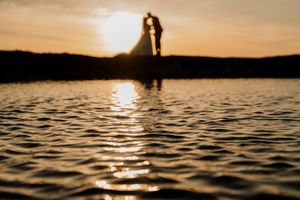 Silhouette of bride and groom reflected in calm water during a breathtaking golden sunset.