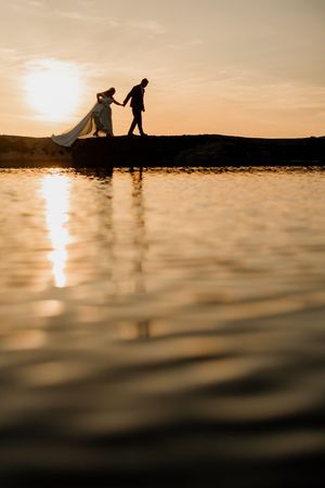 Silhouette of bride and groom walking hand in hand by the water during a golden sunset.