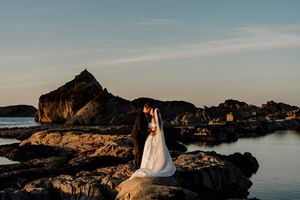 Bride and groom posing on coastal rocks at golden hour with dramatic ocean backdrop.