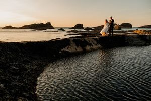 Bride and groom standing on coastal rocks at golden sunset with dramatic ocean and island backdrop.