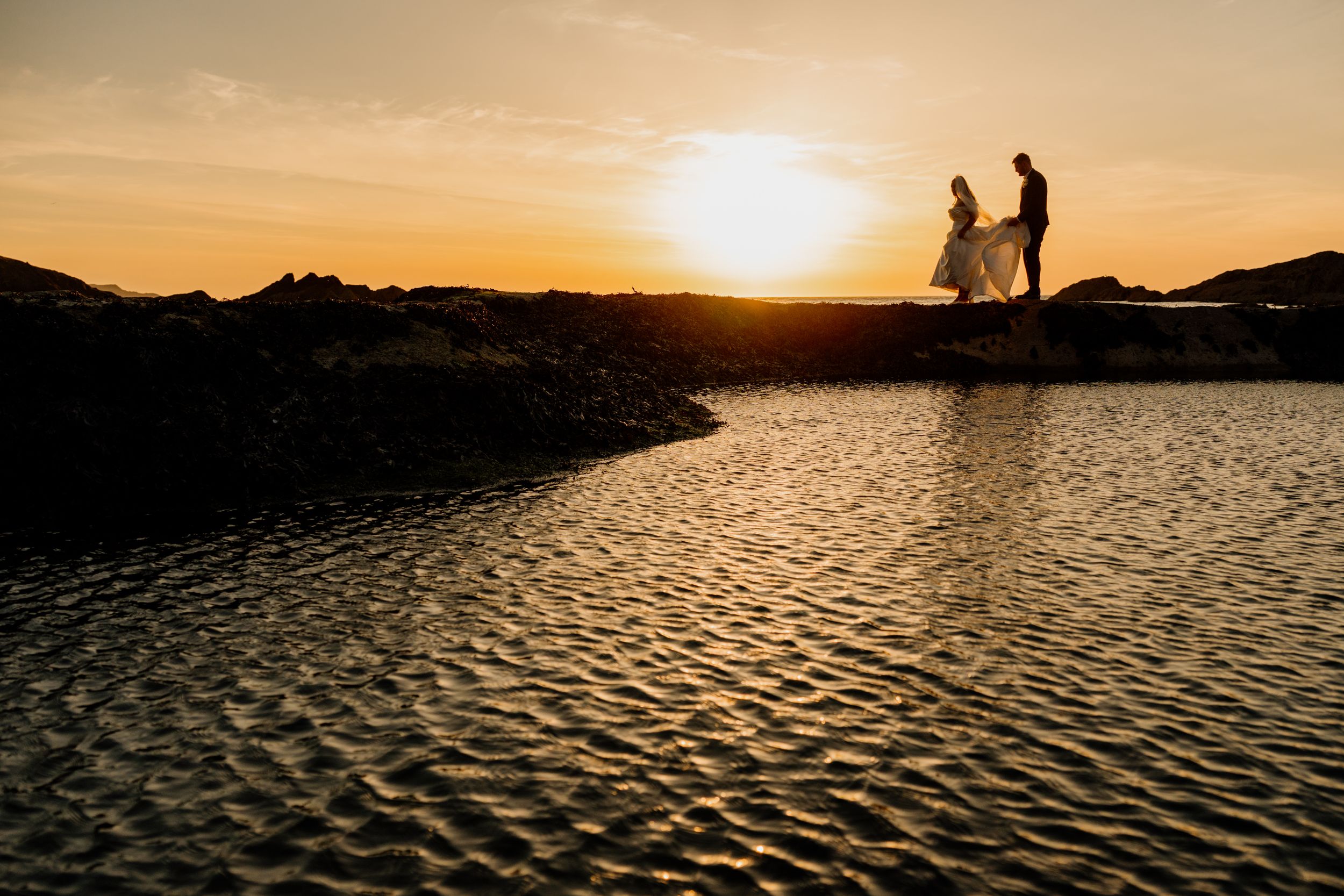 Silhouette of bride and groom standing on rocks beside water during a vivid orange sunset.