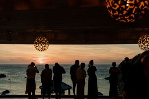 Wedding guests silhouetted against a stunning ocean sunset viewed from a decorated venue terrace.