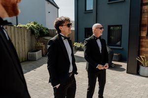 Groomsmen in black tuxedos with bow ties standing outdoors in sunlight before a wedding ceremony.