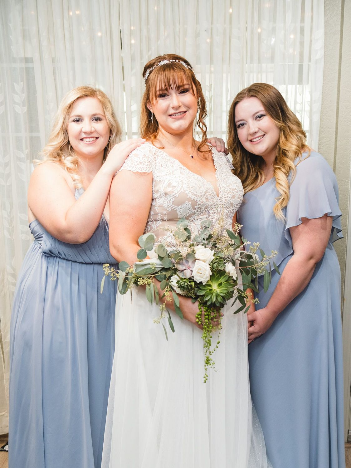 Bride in white lace gown holds bouquet flanked by two bridesmaids in dusty blue dresses at wedding.
