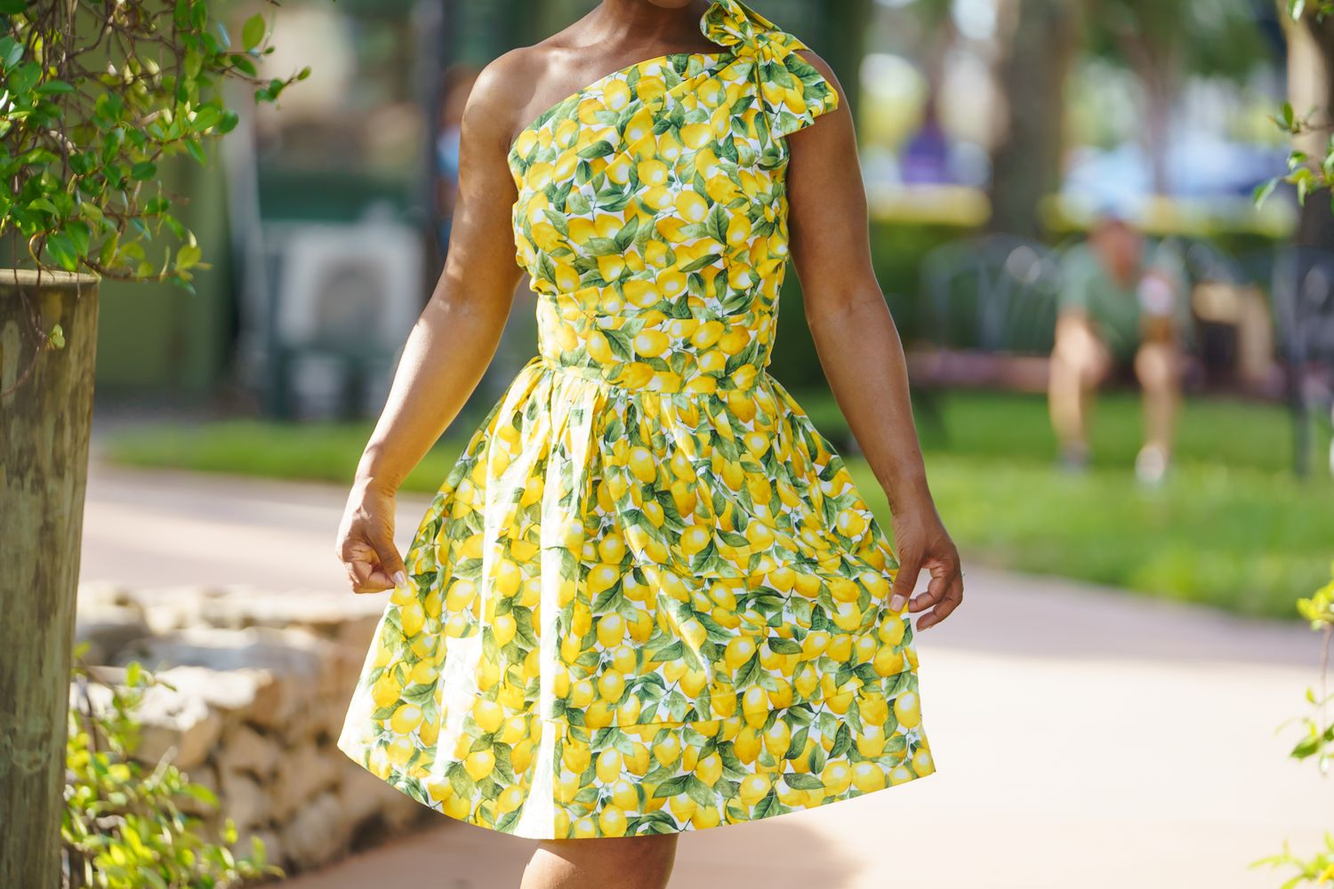 Woman wearing a yellow floral one-shoulder dress walking outdoors on a sunny day.