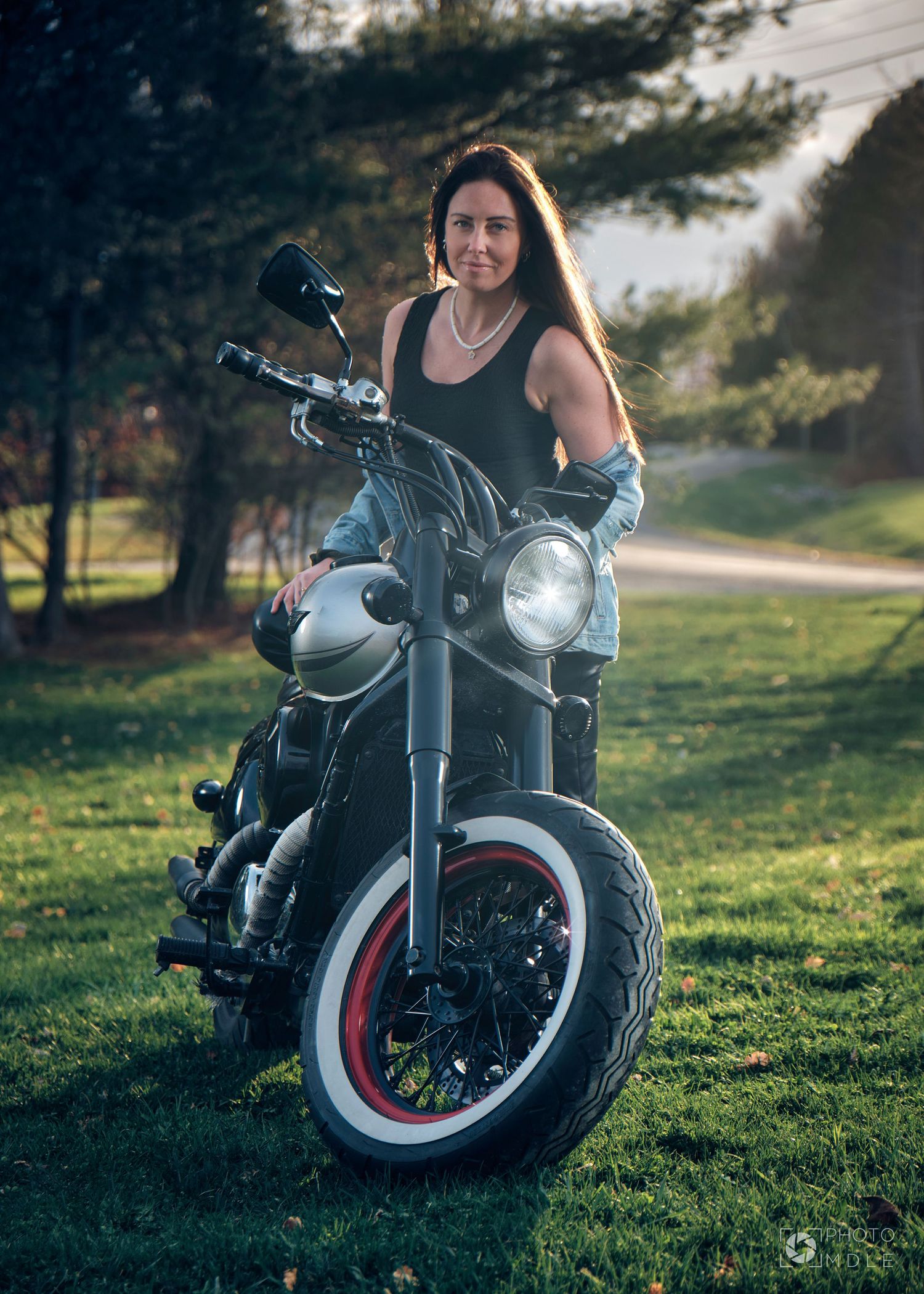 Une jeune femme debout derrière sa moto, éclairée par les rayons du soleil couchant.