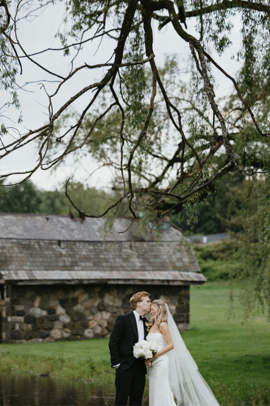 A bride and groom share a kiss outdoors near a rustic stone building, surrounded by lush greenery and overhanging tree branches.
