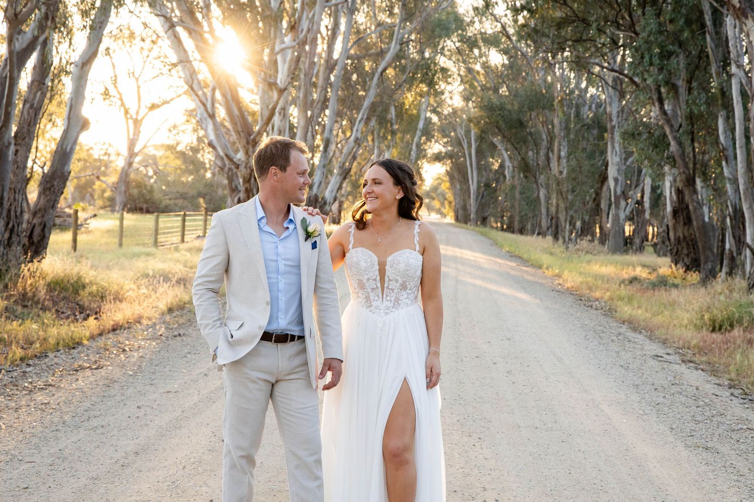 Bride and groom walking hand in hand on a sunlit dirt road surrounded by tall eucalyptus trees at golden hour.