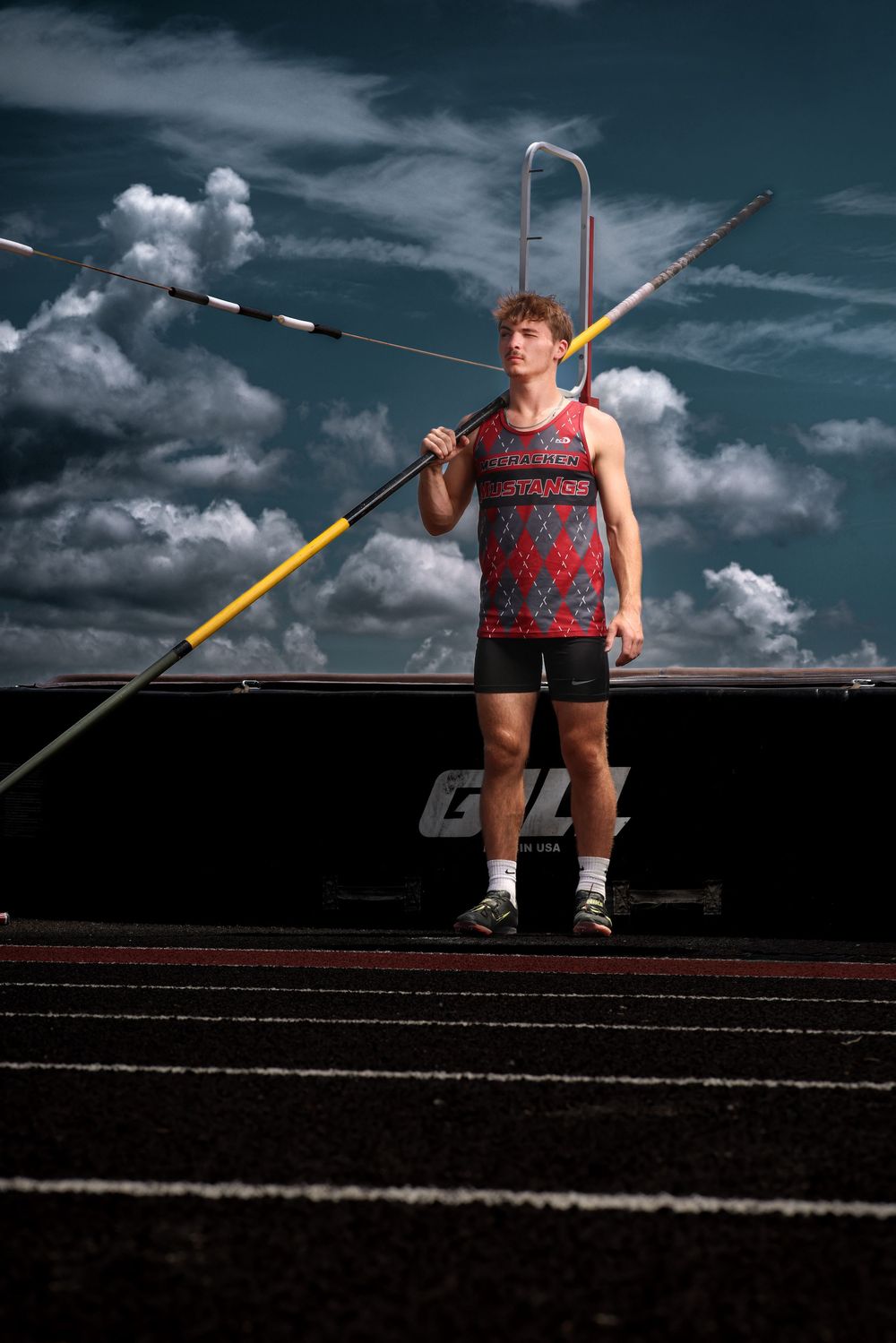 Young male javelin thrower in red athletic uniform poses on track under dramatic cloudy sky.